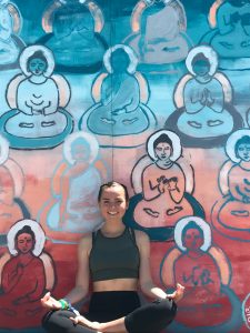 A woman in athletic clothing sits cross-legged and smiling in front of a colorful mural featuring multiple stylized Buddha figures in meditative poses.