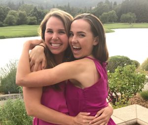 Two young women wearing matching sleeveless magenta dresses are hugging and smiling outdoors by a lake, with greenery and trees in the background.