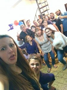 A group of people of various ages pose indoors, making funny faces and playful expressions. One woman holds a small white dog. Balloons are visible in the background, suggesting a celebration or party.