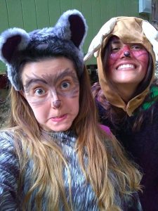 Two people smiling and posing for a selfie in animal costumes; one is dressed as a gray furry animal with painted face, and the other in a brown costume with floppy ears and a painted face.