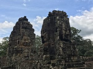 Ancient stone towers at Angkor Thom in Cambodia, featuring large carved faces with serene expressions, set against a partly cloudy blue sky and surrounded by green trees.