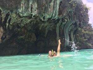 Two people in snorkel gear smile and pose in turquoise water near a large rocky cliff with overhanging, mossy formations and lush greenery above. One person raises an arm, splashing water.