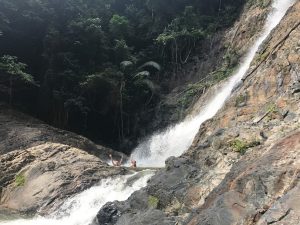 Two people stand on rocks at the base of a waterfall, raising their arms, surrounded by lush green jungle and steep rocky cliffs. Water cascades down from the right side of the image.