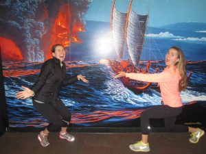 Two women pose playfully in front of a mural showing a historic sailing canoe on the ocean with erupting volcanoes and lava in the background. They mimic swimming or paddling motions, smiling and having fun.