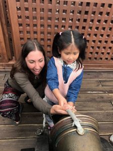 An adult woman kneels beside a young girl as they hold and pull a thick rope together on a wooden deck, with a wooden lattice fence in the background. Both are smiling and focused on the task.