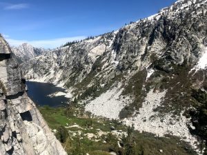 A scenic mountain landscape features steep, rocky slopes partially covered in snow, a blue alpine lake, and patches of greenery under a clear blue sky.