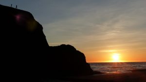Silhouette of two people standing atop a rocky cliff overlooking the ocean at sunset, with the sun low on the horizon and the sky tinged with orange and yellow hues.