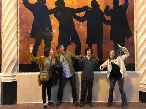 Four people stand in front of a wall mural showing silhouettes of actors in dramatic poses. The group mimics the poses of the silhouettes behind them, smiling and having fun.