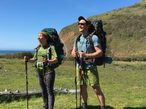 Two hikers with large backpacks and trekking poles stand on a grassy trail, smiling and looking up. Behind them are green hills, blue sky, and the ocean in the distance.