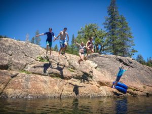 Five people are jumping off a rocky ledge into a lake on a sunny day, with trees and a clear blue sky in the background. One person is holding a blue inner tube while others are mid-air.