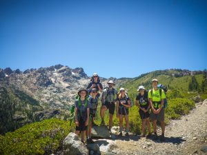 A group of seven hikers stands on a rocky mountain trail, smiling at the camera with backpacks and hats, against a backdrop of rugged, tree-covered peaks and a clear blue sky.