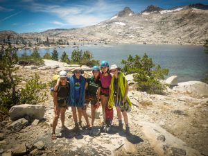 Five people wearing climbing gear and helmets stand together smiling on a rocky lakeshore, with mountains, trees, and a clear blue sky in the background. Coiled ropes are visible around their shoulders.
