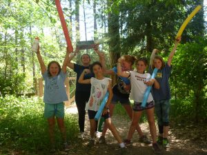 Six children stand on a forest path holding colorful foam pool noodles and smiling, with trees and sunlight in the background, appearing joyful and energetic as if at summer camp.
