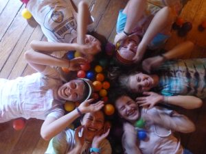 Six smiling people lie in a circle on a wooden floor, heads together, surrounded by colorful plastic balls, with their hands raised playfully near their faces.
