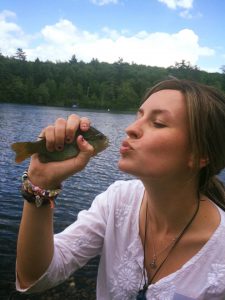 A woman in a white shirt holds a small fish near her lips as if giving it a kiss, standing by a lake with a forested shoreline in the background under a partly cloudy sky.