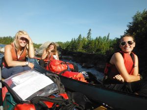 Three women wearing life jackets sit in a canoe surrounded by gear. They are smiling and enjoying a sunny day; trees and rocks are visible along the shoreline in the background.