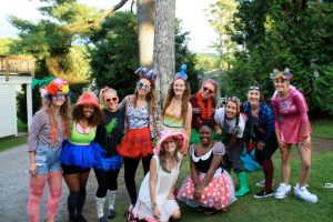 A group of eleven people dressed in colorful, quirky costumes and accessories, standing and kneeling outdoors on grass, smiling and posing together in front of trees and a white building.