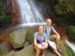 Two people sit on large rocks in front of a waterfall in a lush, green forest. Both are smiling at the camera, and one holds a hat. The scene is bright and natural, with water flowing behind them.