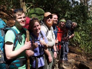A group of seven smiling hikers with backpacks stand in a sunlit forest, posing closely together on a trail among trees and greenery. Some are holding hiking sticks and wearing sunglasses or headbands.