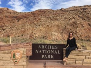 A woman in black sits and smiles next to the entrance sign for Arches National Park, with rocky red cliffs and blue sky in the background.