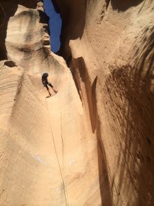A person with a backpack rappels down a steep, narrow, sunlit sandstone canyon wall with a rope, surrounded by tall, smooth rock formations and a patch of blue sky above.