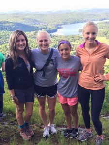 Four young women stand together smiling on a grassy hilltop with a scenic view of green forests and a lake in the background on a sunny day.