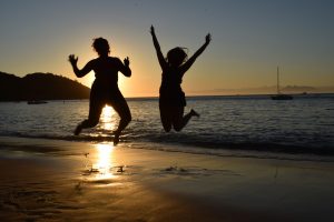 Two people are jumping in the air on a beach at sunset, their silhouettes visible against the golden sky and water. A sailboat floats in the distance, and hills line the horizon.