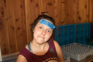 A young woman with light brown hair in a bun makes a funny face. She has a blue sticky note on her forehead with writing on it. The background features wooden walls and blue crates.