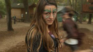 A girl with long brown hair and green face paint in leaf patterns stands outdoors, looking at the camera. Blurred people and wooden cabins are visible in the forested background.