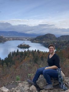 Dani sitting on a rock overlooking Lake Bled which has a church on a island in the middle and mountains in the background.