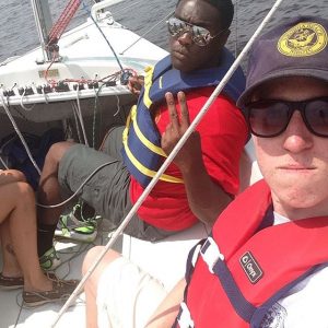 Two people wearing life jackets sit on a sailboat. One person in a red shirt makes a peace sign, and the other, wearing a cap and sunglasses, takes a selfie. Part of another person’s legs are visible on the left.