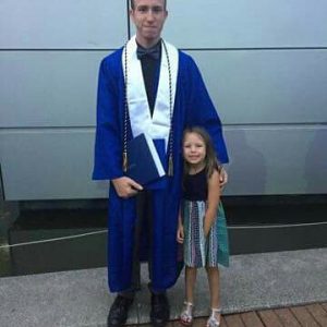 A young man in a blue graduation gown and cap stands next to a smiling young girl in a colorful dress, both posing for a photo outdoors in front of a gray wall.