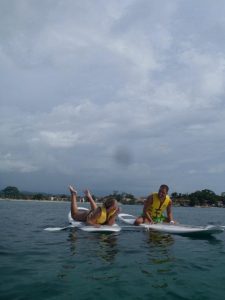 Two people wearing yellow life jackets sit on paddleboards in the water; one laughs while falling back, and the other smiles, with a cloudy sky and shoreline visible in the background.