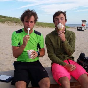 Two young men sit on a low wall at a sandy beach eating ice cream. One wears a neon green shirt and holds a cup with a spoon, while the other wears a green jacket and pink shorts, holding an ice cream cone. The ocean is in the background.