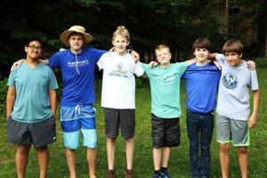 Six boys stand arm-in-arm in a row outdoors on grass, smiling at the camera. They are dressed casually in t-shirts and shorts. Tall trees and greenery are visible in the background.