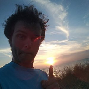 A person with tousled hair points toward a sunset over the ocean, with grasses in the foreground and wispy clouds in the sky.