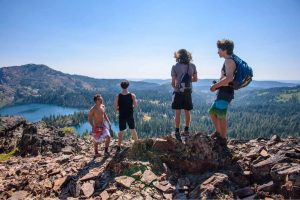 Four people stand on rocky terrain overlooking a scenic landscape with a lake, forest, and distant mountains under a clear blue sky. Two have backpacks and all are dressed in summer clothes.