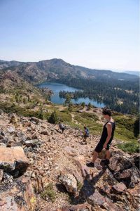 Three hikers descend a rocky trail overlooking a forested mountain landscape with a blue lake nestled among trees under a clear sky.