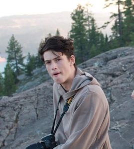A young person in a tan hoodie sits on a rocky hill with a camera around their neck, surrounded by pine trees and distant mountains in the background.