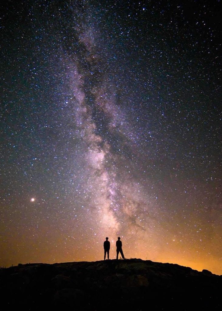 Two people stand on a hilltop, silhouetted against a vibrant, star-filled night sky with the Milky Way galaxy clearly visible above them.