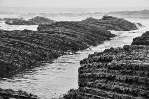 Black and white photo of jagged, layered rock formations along a shoreline, with ocean waves breaking in the background and calm tide pools in the foreground.