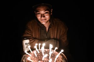 A person wearing a jacket and cap smiles in dim light, sitting behind a birthday cake with lit candles, and a beer can labeled Modelo is on the table to the left. The background is dark.