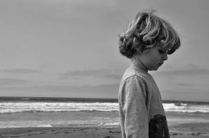 Black and white photo of a young boy with wavy hair standing sideways on a beach, looking down thoughtfully, with waves and a cloudy sky in the background.