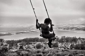 A child on a swing soars high above a scenic landscape, with a body of water, distant mountains, and a town visible below. The image is in black and white.