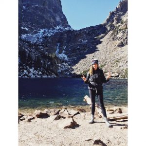 A woman wearing outdoor gear stands smiling on a rocky shore in front of a deep blue mountain lake, with tall, snow-dusted cliffs and pine trees in the background under bright sunlight.