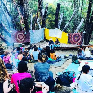 A group of people sit on blankets and tarps outdoors, facing a small stage with two people talking. The stage is decorated with colorful fabrics and surrounded by trees. It appears to be a camp or outdoor gathering.