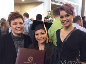 Three women smile at a graduation ceremony. The woman in the center wears a cap and gown and holds a diploma. The two women beside her are casually dressed. People and balloons appear in the background.