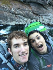 Two young people with backpacks smile and make playful faces while taking a selfie outdoors near a rocky river. They appear to be hiking or camping, with water and rocks visible in the background.
