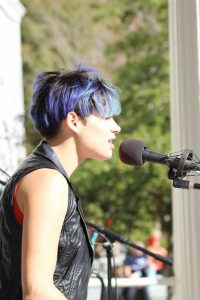 A person with short blue and purple hair sings into a microphone outdoors, wearing a black sleeveless vest. The background is blurred with greenery and sunlight.