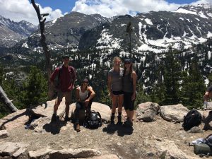 Four hikers pose on a rocky outcrop with backpacks, surrounded by pine trees and snowy mountain peaks under a partly cloudy sky.